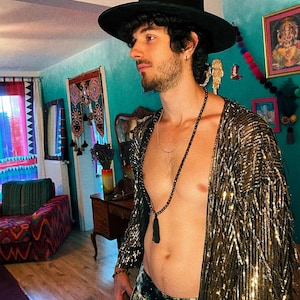 May include: A black wide-brimmed hat, a long gold-toned sequined open-front cardigan with fringe detailing, and a long black beaded necklace with a tassel pendant. The cardigan features a metallic finish that reflects light across its textured surface.