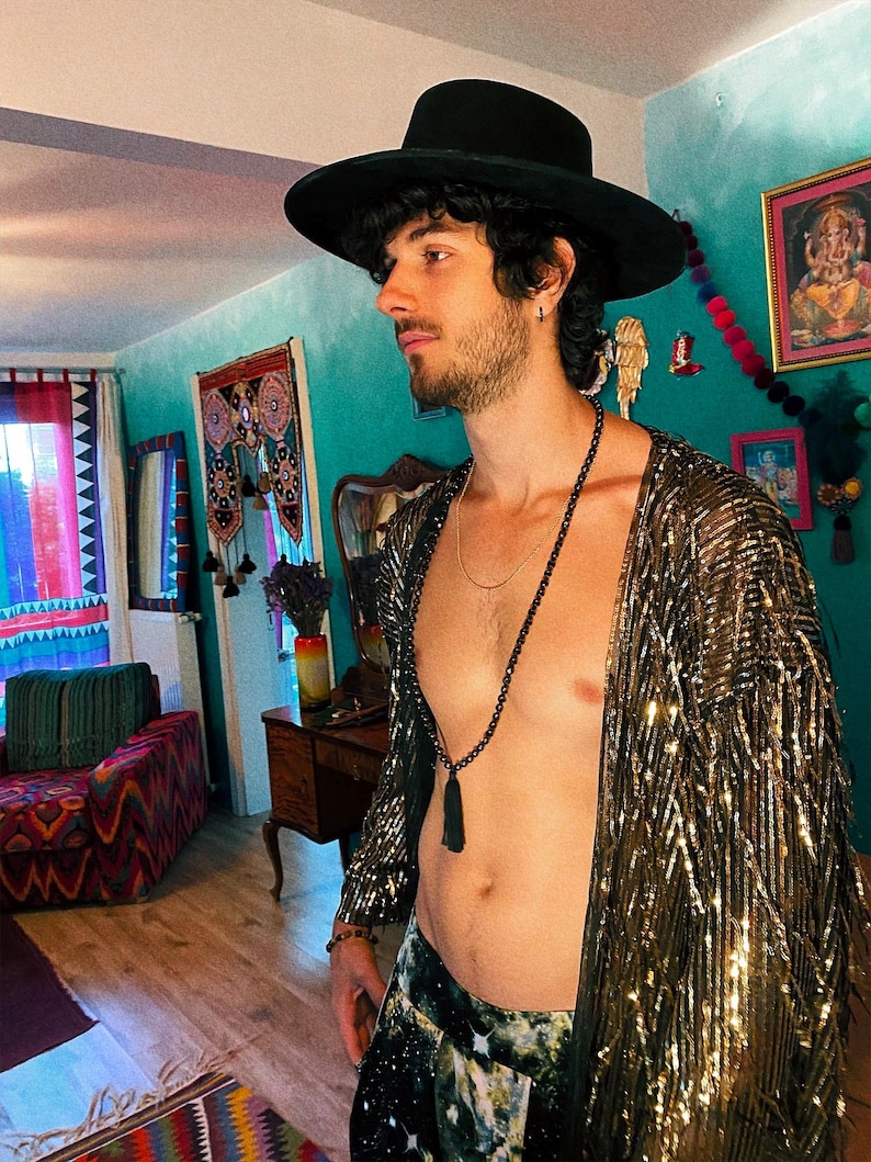 May include: A black wide-brimmed hat, a long gold-toned sequined open-front cardigan with fringe detailing, and a long black beaded necklace with a tassel pendant. The cardigan features a metallic finish that reflects light across its textured surface.