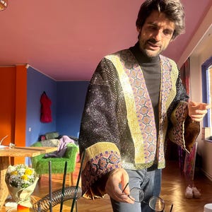 May include: A black open-front jacket featuring wide sleeves and gold-toned trim with intricate geometric patterns. The garment has a shimmering, textured surface and is worn over a black turtleneck sweater and dark trousers, paired with round glasses.