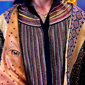 May include: A multi-colored striped shirt featuring a black collar and sheer mesh panels at the neckline with horizontal embroidery. The garment includes gold-toned satin lapels printed with black star patterns and eye illustrations, layered over a black base.
