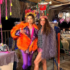 May include: A bright orange faux fur jacket with a purple sequined lapel, worn with purple leggings and black boots. Beside it, a grey shaggy faux fur jacket is paired with a black and white patterned shirt, pink shorts, and grey platform sandals.