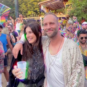 May include: A person wearing a black sleeveless top with intricate sequin and fringe detailing, paired with a light-colored sequined cardigan. The outfit includes a white undershirt, layered necklaces, and a clear plastic cup held in one hand.