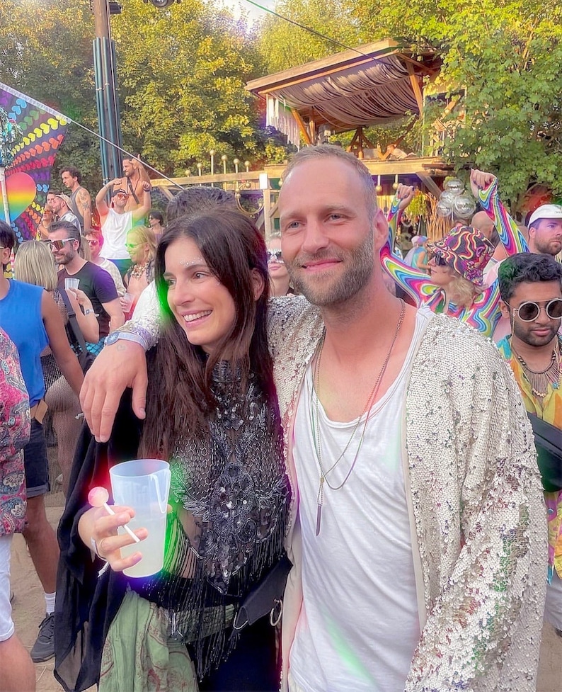 May include: A person wearing a black sleeveless top with intricate sequin and fringe detailing, paired with a light-colored sequined cardigan. The outfit includes a white undershirt, layered necklaces, and a clear plastic cup held in one hand.