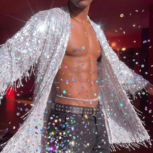 May include: A long-sleeved silver sequined duster jacket with fringe trim, worn over black trousers with a studded belt and a thin rhinestone waist chain. The garment features a shimmering, reflective surface that catches light, creating a sparkling effect.