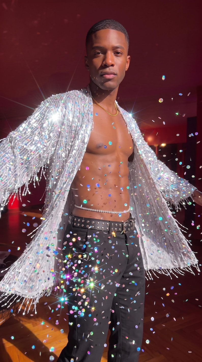 May include: A long-sleeved silver sequined duster jacket with fringe trim, worn over black trousers with a studded belt and a thin rhinestone waist chain. The garment features a shimmering, reflective surface that catches light, creating a sparkling effect.