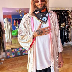 May include: A hooded robe covered in shimmering champagne-colored sequins with a pink inner lining. The robe is worn over a plain white t-shirt, black patterned shorts, and a black bandana featuring a colorful geometric and cube print design.