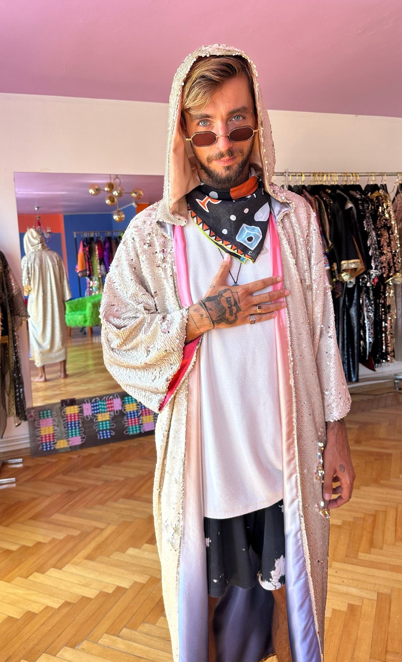 May include: A hooded robe covered in shimmering champagne-colored sequins with a pink inner lining. The robe is worn over a plain white t-shirt, black patterned shorts, and a black bandana featuring a colorful geometric and cube print design.