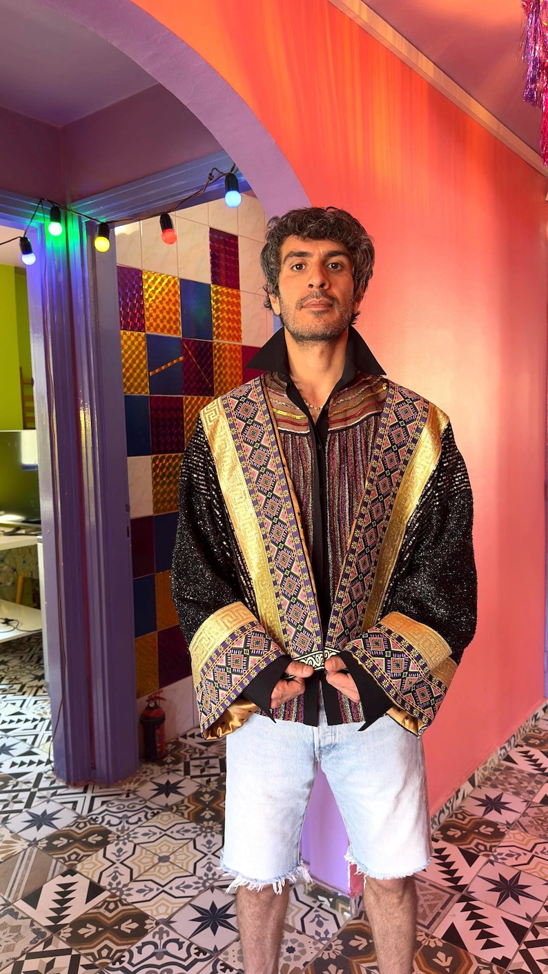 May include: A long-sleeved jacket featuring black fabric with a shimmering texture, accented by wide gold bands and intricate geometric patterns in purple, blue, and orange. The garment has a high collar, wide cuffs, and a structured open front design.
