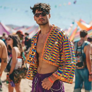Puede incluir: Una chaqueta corta abierta con un patrón de rayas horizontales de arcoíris vibrantes y un borde geométrico en contraste. La prenda se combina con pantalones morados que incluyen un detalle de cadena plateada en la cintura.