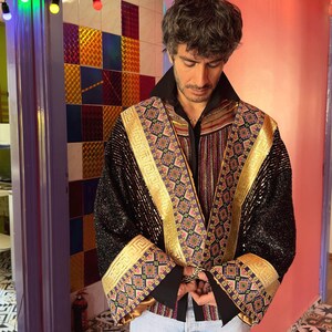 May include: A black jacket featuring gold metallic trim and intricate multi-colored geometric embroidery along the lapels and wide cuffs. The garment is worn over a black collared shirt and light blue denim shorts, showcasing a textured finish.