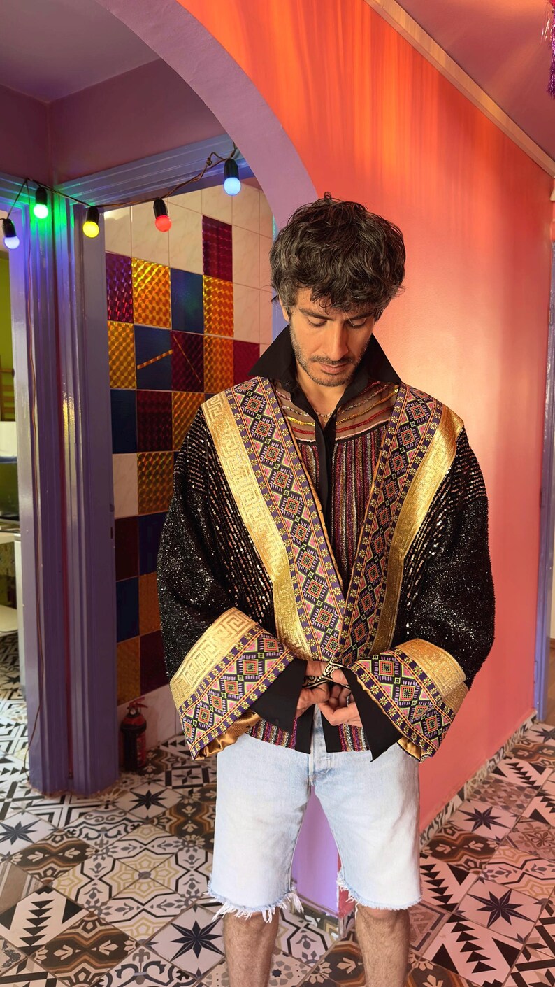 May include: A black jacket featuring gold metallic trim and intricate multi-colored geometric embroidery along the lapels and wide cuffs. The garment is worn over a black collared shirt and light blue denim shorts, showcasing a textured finish.