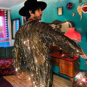 May include: A long-sleeved jacket covered in metallic gold fringe that creates a shimmering effect. The garment features a high-neck collar and a straight, loose fit that extends to the mid-thigh. The fringe is dense and reflective.