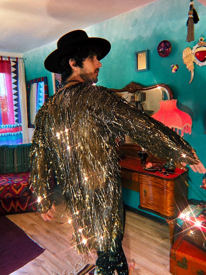 May include: A long-sleeved jacket covered in metallic gold fringe that creates a shimmering effect. The garment features a high-neck collar and a straight, loose fit that extends to the mid-thigh. The fringe is dense and reflective.