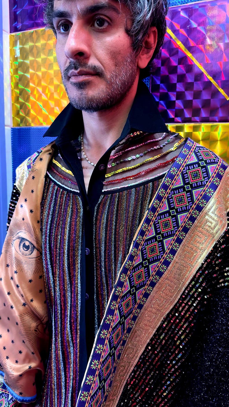 May include: A black button-down shirt featuring a vertical metallic striped panel, a sheer yoke with horizontal beaded rows, and a wide satin shoulder panel printed with a large eye and star motif, accented by a decorative blue and gold geometric trim.