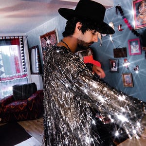 May include: A long-sleeved shirt covered in shimmering silver sequins and fringe, paired with a wide-brimmed black hat. The garment features a dense layer of metallic silver fringe that catches the light, creating a reflective and textured appearance.