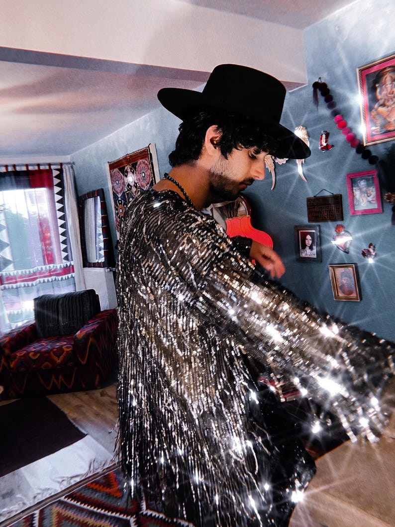 May include: A long-sleeved shirt covered in shimmering silver sequins and fringe, paired with a wide-brimmed black hat. The garment features a dense layer of metallic silver fringe that catches the light, creating a reflective and textured appearance.