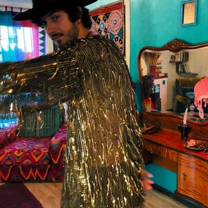 May include: A long-sleeved jacket covered in metallic gold fringe that hangs down from the shoulders and sleeves. The garment is worn with a wide-brimmed black hat. The fringe creates a reflective, textured surface that catches light across the entire piece.