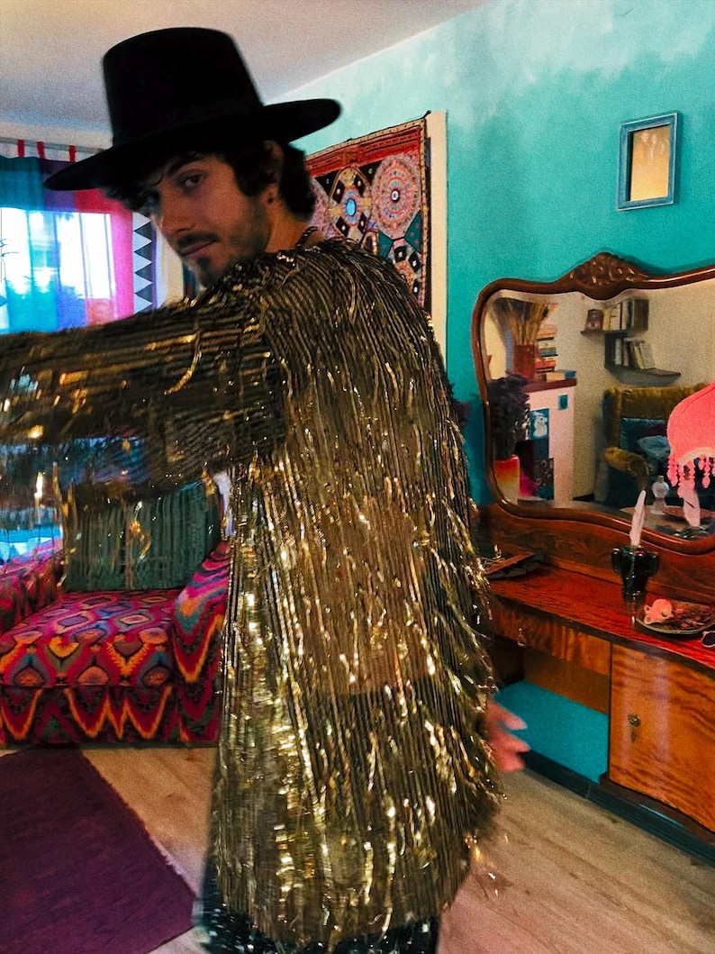 May include: A long-sleeved jacket covered in metallic gold fringe that hangs down from the shoulders and sleeves. The garment is worn with a wide-brimmed black hat. The fringe creates a reflective, textured surface that catches light across the entire piece.