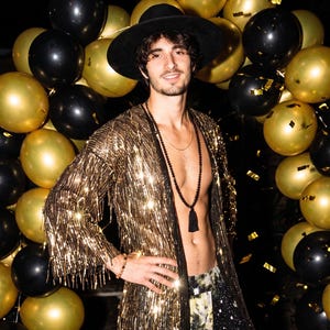 May include: A long-sleeved gold sequined duster jacket with fringe trim, worn with a black wide-brimmed hat, a long beaded necklace with a black tassel, and dark patterned trousers, set against a backdrop of black and gold balloons.