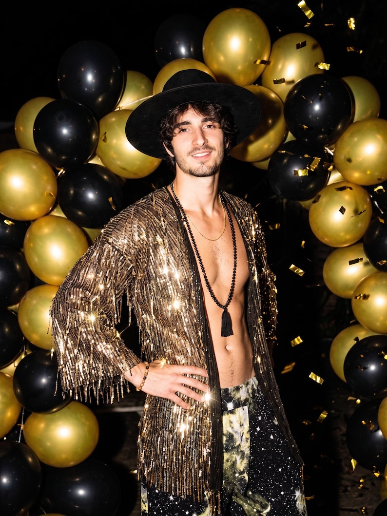 May include: A long-sleeved gold sequined duster jacket with fringe trim, worn with a black wide-brimmed hat, a long beaded necklace with a black tassel, and dark patterned trousers, set against a backdrop of black and gold balloons.