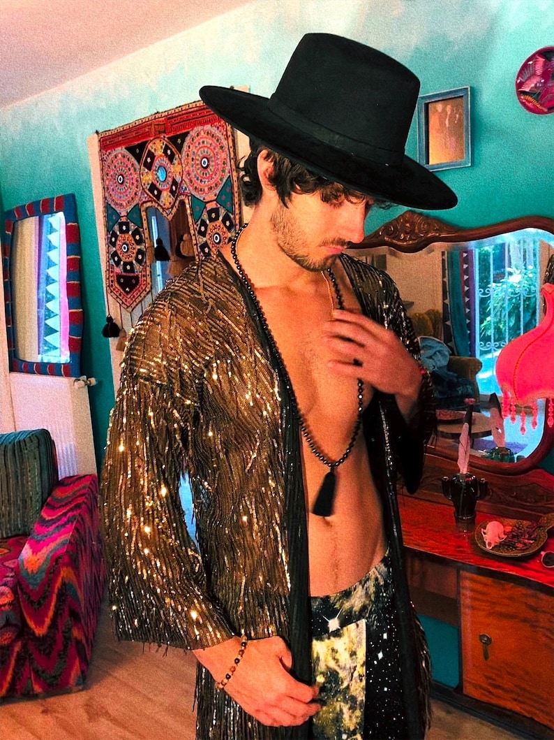 May include: A black wide-brimmed hat, a long-sleeved gold sequined duster with fringe detailing, and a long black beaded necklace with a tassel. The garment is worn open over the torso, paired with patterned trousers and a beaded wrist bracelet.