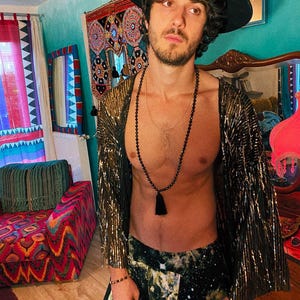 May include: A black wide-brimmed hat, a metallic gold textured open-front kimono with wide sleeves, a long black beaded necklace with a tassel, and dark trousers featuring a white and green galaxy-inspired print with a star-like pattern.