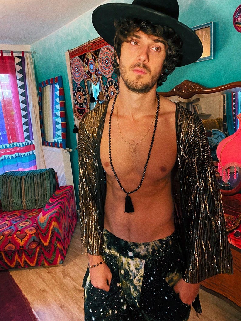 May include: A black wide-brimmed hat, a metallic gold textured open-front kimono with wide sleeves, a long black beaded necklace with a tassel, and dark trousers featuring a white and green galaxy-inspired print with a star-like pattern.
