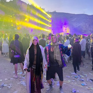 May include: Two individuals wearing layered festival attire, including a long shimmering silver robe, a dark scarf, a deep red skirt with decorative tassels, a dark tank top, black shorts, a blue sequined jacket, and chunky platform boots.