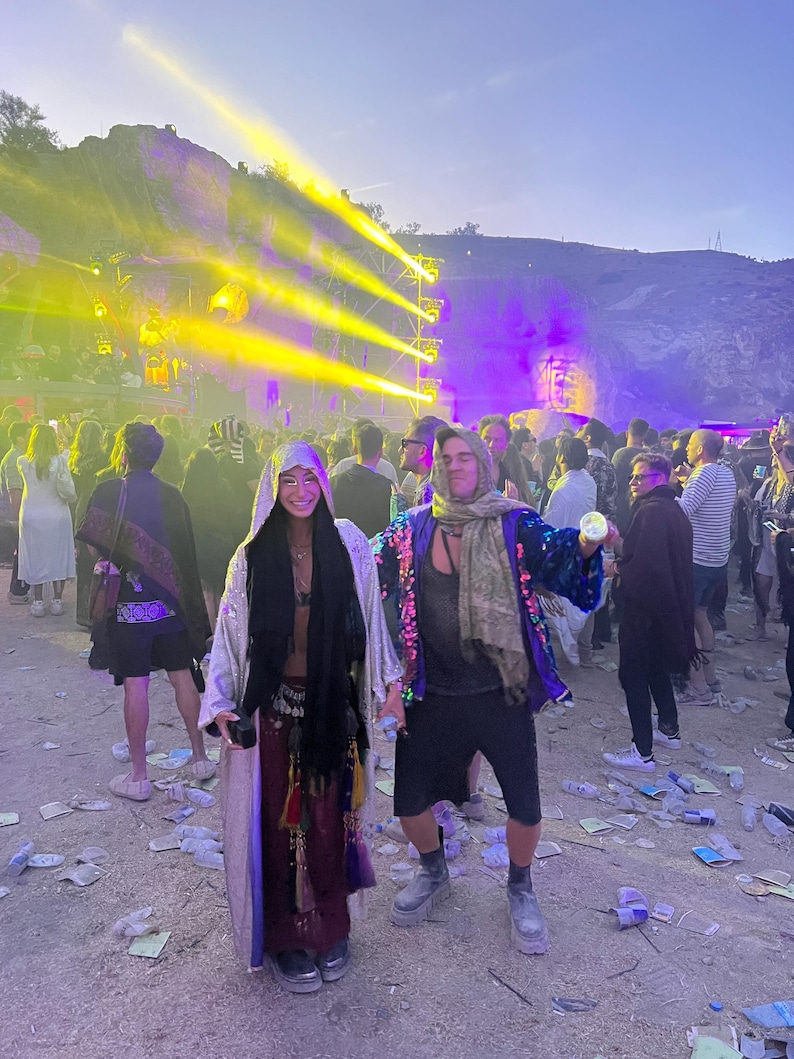 May include: Two individuals wearing layered festival attire, including a long shimmering silver robe, a dark scarf, a deep red skirt with decorative tassels, a dark tank top, black shorts, a blue sequined jacket, and chunky platform boots.