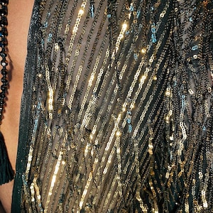May include: A dark green sheer mesh garment featuring vertical rows of shimmering gold sequins arranged in a diagonal pattern. The fabric is lightweight and semi-transparent, with the metallic sequins catching the light to create a textured effect.