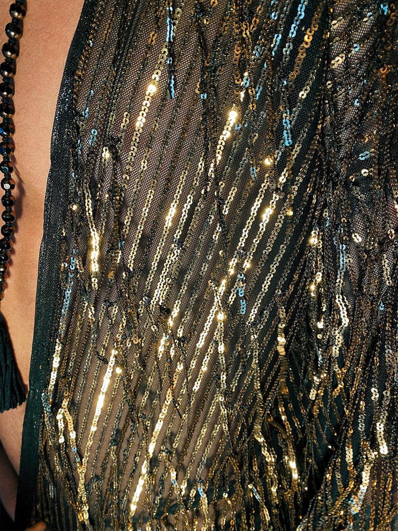 May include: A dark green sheer mesh garment featuring vertical rows of shimmering gold sequins arranged in a diagonal pattern. The fabric is lightweight and semi-transparent, with the metallic sequins catching the light to create a textured effect.