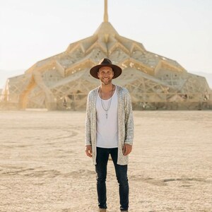 May include: A brown wide-brimmed hat, a silver sequined long cardigan, a plain white tank top, dark blue denim jeans, and tan lace-up boots. The outfit includes a layered silver necklace with a pendant, worn in an outdoor desert environment.