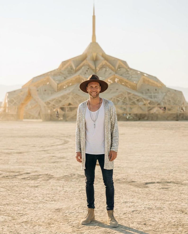 May include: A brown wide-brimmed hat, a silver sequined long cardigan, a plain white tank top, dark blue denim jeans, and tan lace-up boots. The outfit includes a layered silver necklace with a pendant, worn in an outdoor desert environment.