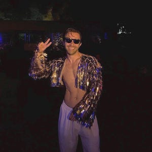 May include: A cropped jacket with gold and blue fringe is worn by a person. The jacket has long sleeves and is open in the front. The person is wearing sunglasses and white pants. The background is dark.