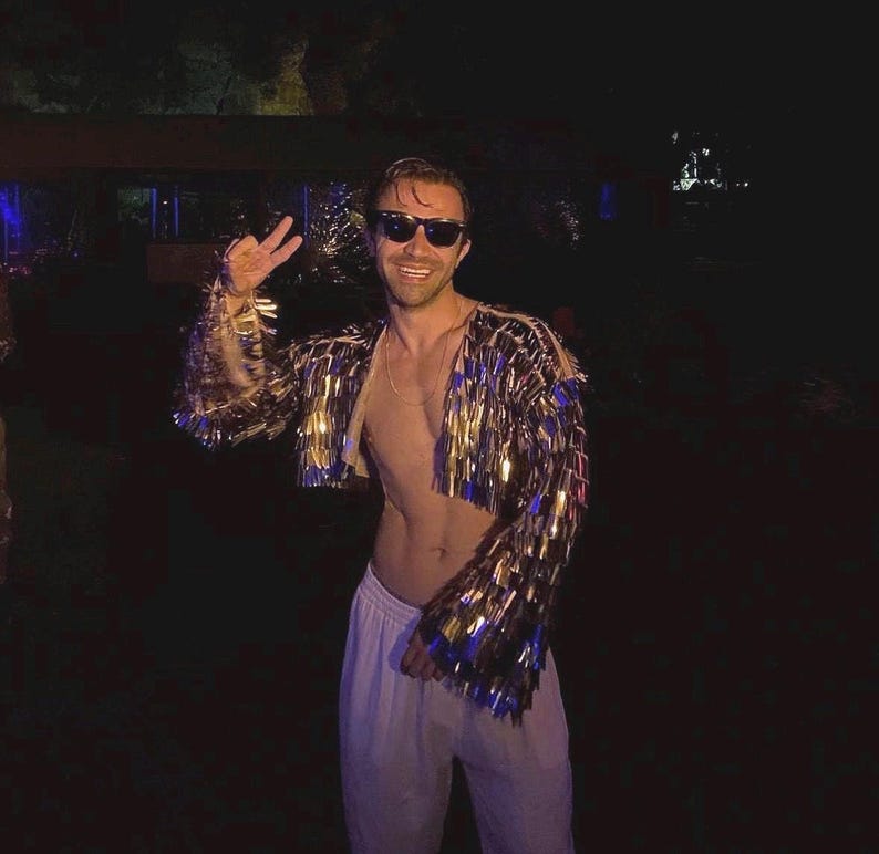 May include: A cropped jacket with gold and blue fringe is worn by a person. The jacket has long sleeves and is open in the front. The person is wearing sunglasses and white pants. The background is dark.
