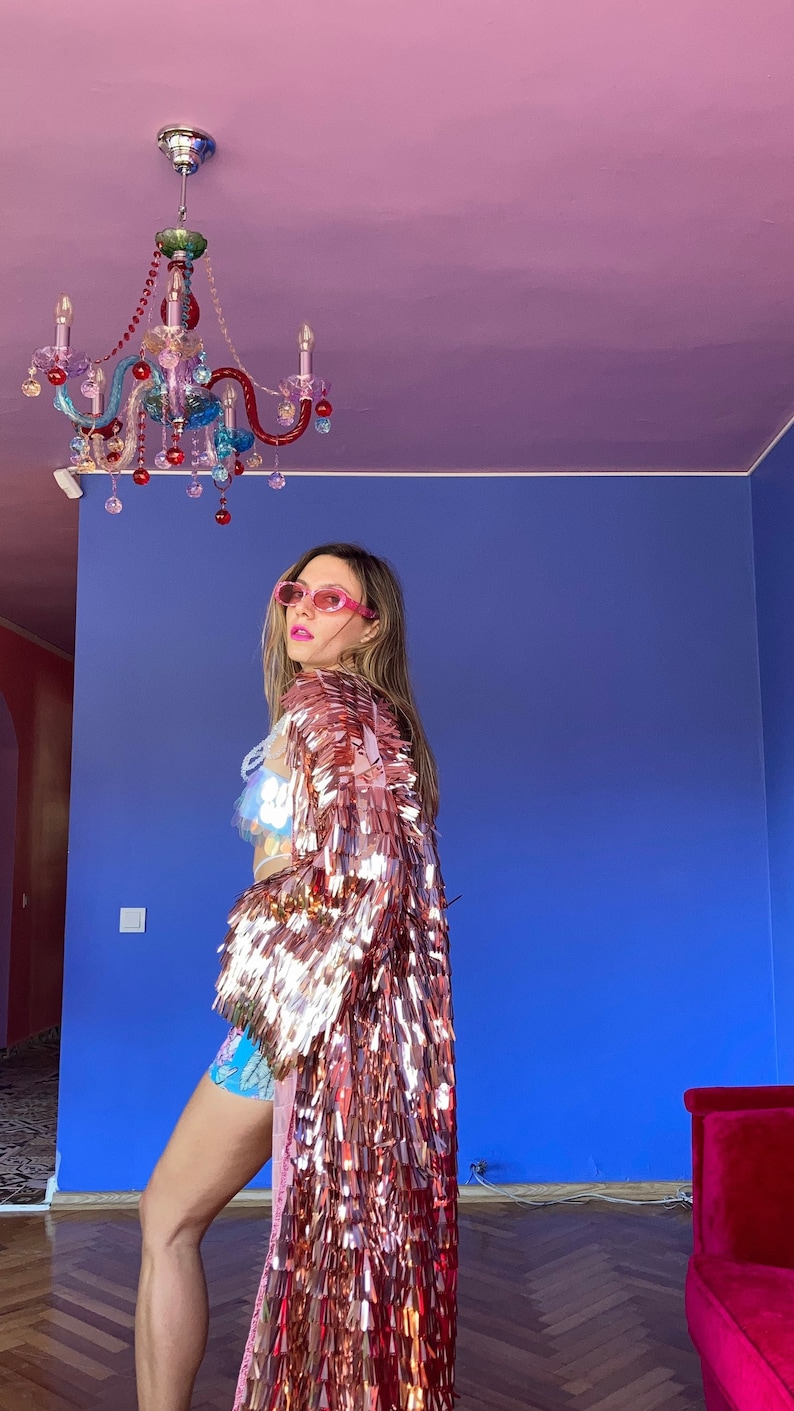 May include: A rose gold sequin jacket with long fringe. The jacket is worn over a sequined top and blue shorts. The background features a blue wall, a pink ceiling, and a colorful chandelier.