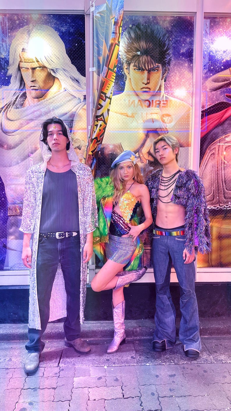 May include: Three people wearing colourful, sparkly clothing pose in front of a shop window with a large graphic of a cartoon character. The person on the left is wearing a silver sequined jacket, black tank top, and blue jeans. The person in the middle is wearing a rainbow sequined top, a green fur jacket, and pink cowboy boots. The person on the right is wearing a purple sequined jacket, a beaded necklace, and blue jeans.