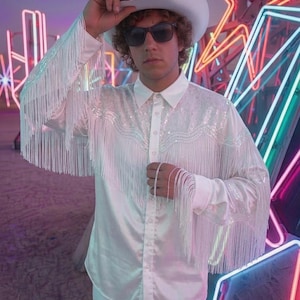 May include: A white long-sleeved button-down shirt featuring a sequined yoke and long fringe detailing on the sleeves and chest. The shirt is paired with a matching white cowboy hat and dark sunglasses, creating a coordinated monochromatic ensemble.