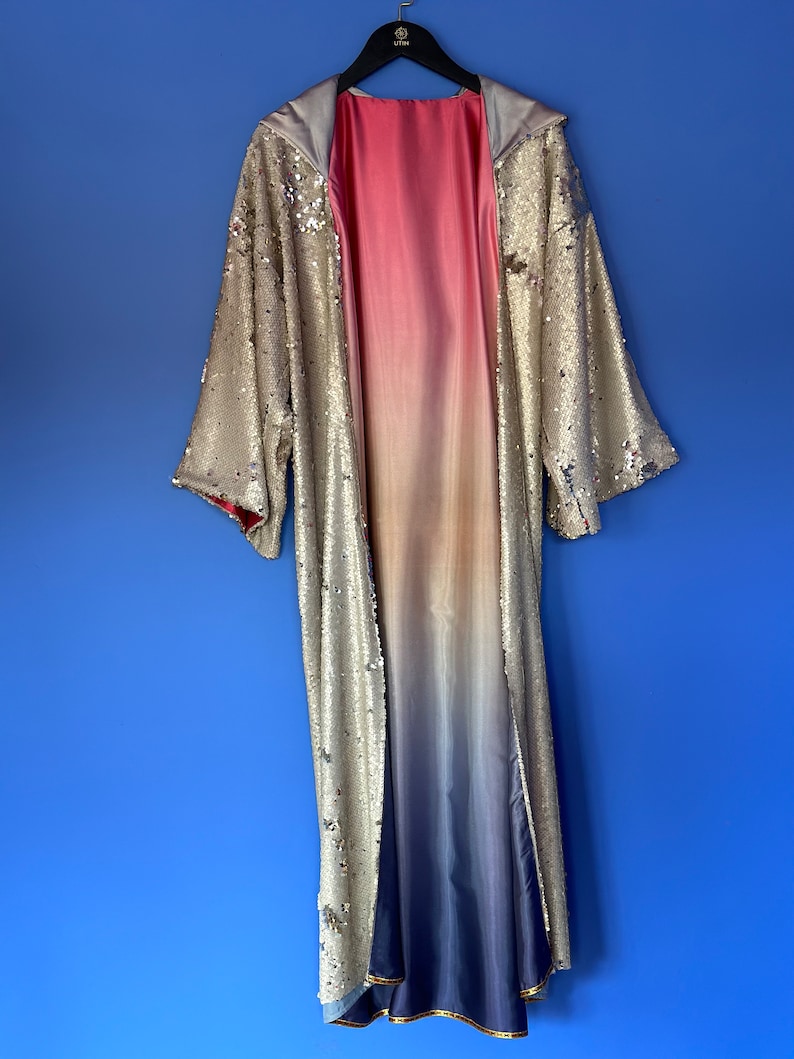 May include: A long, open-front robe featuring a shimmering gold sequined exterior and a smooth, satin-like interior lining that transitions in a gradient from pink at the top to white in the middle and deep blue at the bottom hem.