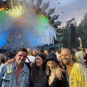 May include: Four individuals stand together at an outdoor music festival. One wears a light blue denim jacket and pink-tinted glasses, another a black embellished top, a third a black bucket hat, and the fourth a bright yellow sequined jacket.