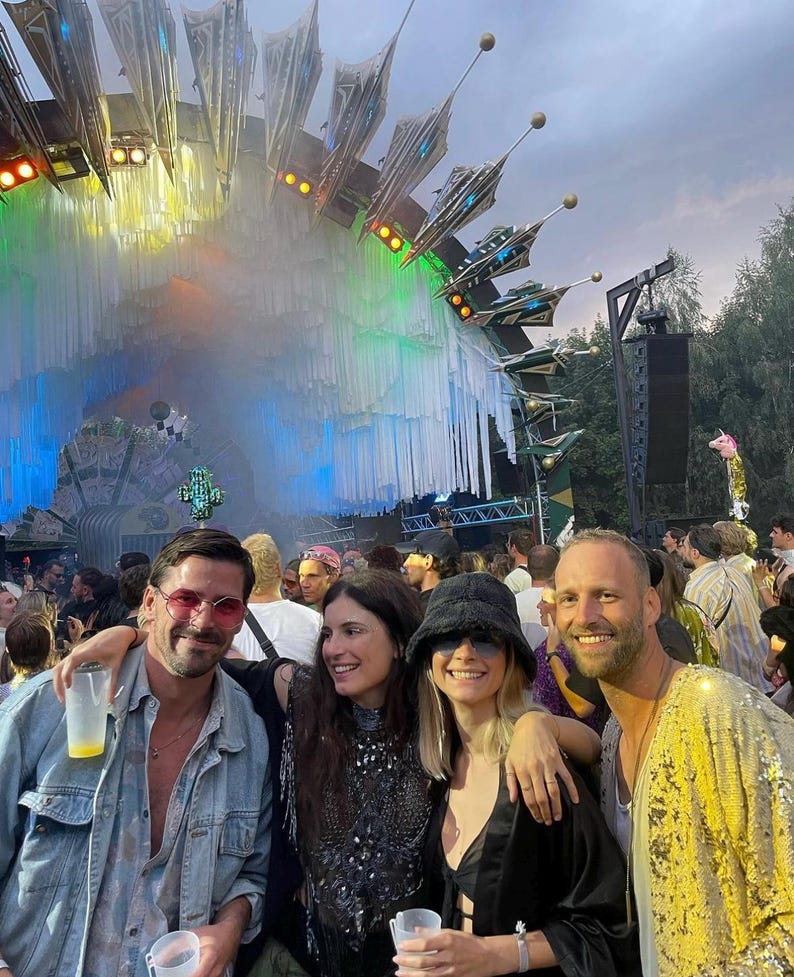 May include: Four individuals stand together at an outdoor music festival. One wears a light blue denim jacket and pink-tinted glasses, another a black embellished top, a third a black bucket hat, and the fourth a bright yellow sequined jacket.
