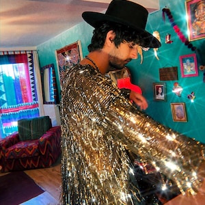 May include: A long-sleeved shirt covered in reflective gold metallic fringe that creates a shimmering effect. The garment features a round neckline and a dense layer of vertical gold strands that move and catch the light, paired with a black hat.