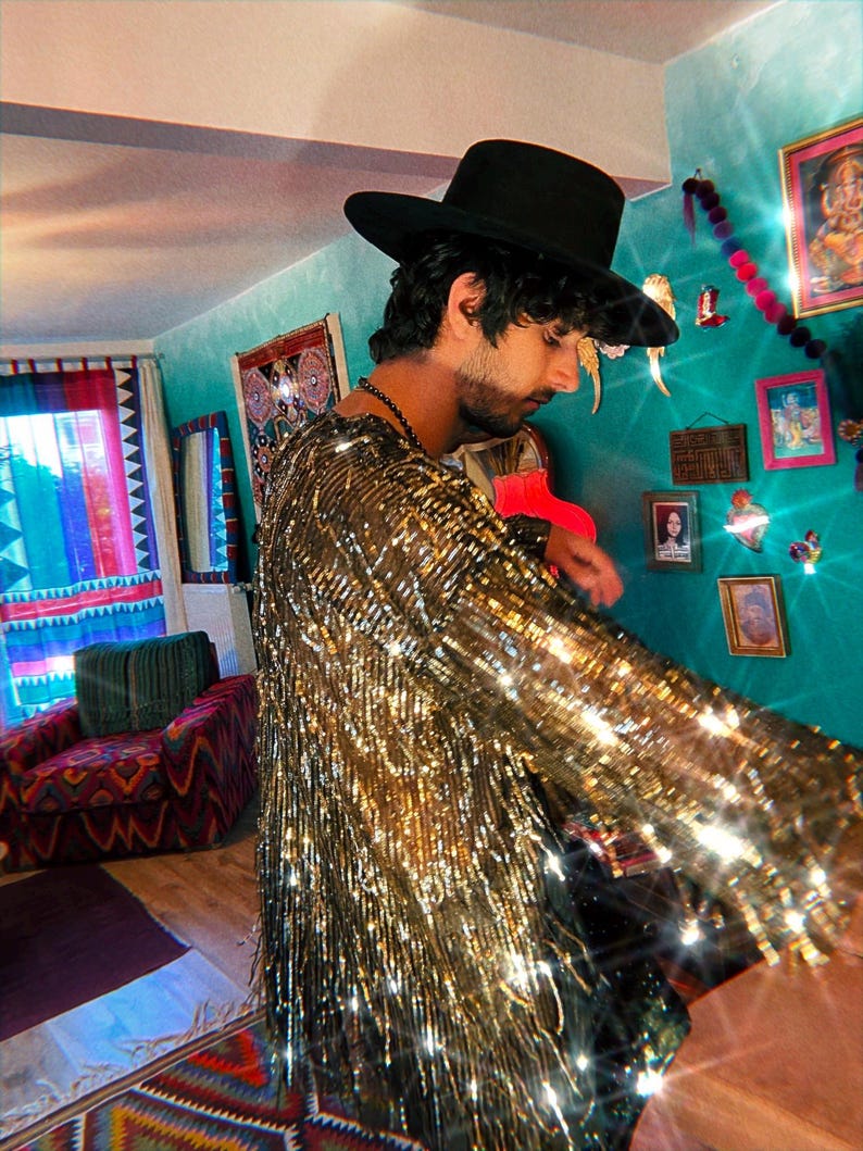 May include: A long-sleeved shirt covered in reflective gold metallic fringe that creates a shimmering effect. The garment features a round neckline and a dense layer of vertical gold strands that move and catch the light, paired with a black hat.