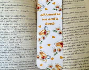 Puede incluir: Marcapáginas blanco con el texto "All I need is tea and a book". Presenta ilustraciones de tazas de té, margaritas y corazones amarillos. El marcapáginas está entre las páginas de un libro.