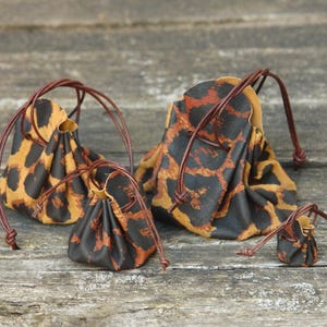 Könnte beinhalten: Vier kleine Beutel mit Kordelzug und Leopardenmuster in Braun-, Schwarz- und Beigetönen. Die Beutel sind aus Stoff und haben braune, lederartige Kordelzüge. Sie sind auf einer verwitterten Holzoberfläche angeordnet.