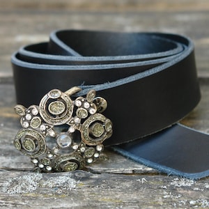 Könnte beinhalten: Ein schwarzer Ledergürtel mit einer dekorativen silberfarbenen Schnalle. Die Schnalle hat ein verziertes Design mit klaren und grün getönten Strasssteinen. Der Gürtel ist aufgerollt und zeigt seine Textur und sein Design.