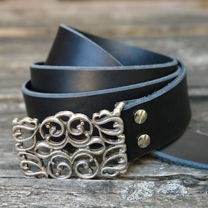 Könnte beinhalten: Ein schwarzer Ledergürtel mit einer dekorativen silberfarbenen Schnalle. Die Schnalle hat ein verziertes, wirbelndes Design. Der Gürtel ist aufgerollt und zeigt seine Textur und Metallbeschläge.