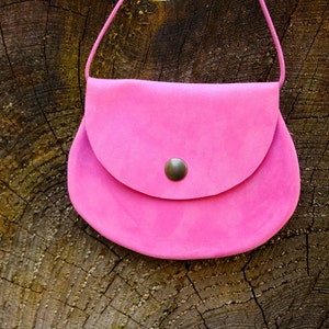 Könnte beinhalten: Eine kleine rosa Wildledertasche mit einer Klappe und einem Metallknopf. Die Tasche hat einen langen Riemen und hängt an einem Baumstumpf.