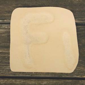 Könnte beinhalten: Ein hellbrauner Lederuntersetzer mit dem Buchstaben "F" auf der Oberfläche. Der Untersetzer hat abgerundete Ecken und eine leicht unregelmäßige Form. Der Hintergrund ist eine dunkle Holzoberfläche.