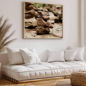 Könnte beinhalten: Ein gerahmter Druck von gestapelten Steinen hängt über einem weißen Futon mit weißen Kissen. Ein geflochtener Hocker steht auf einem Teppich vor dem Futon. Eine hohe Pflanze in einem geflochtenen Topf befindet sich links vom Futon.